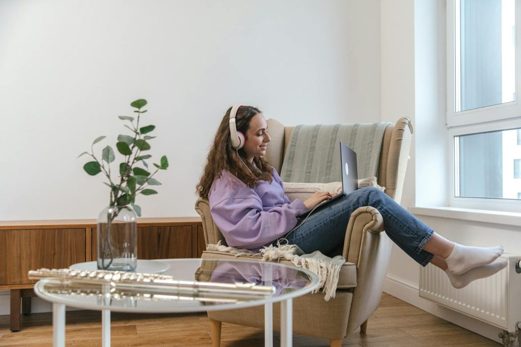 Young woman wearing headphones, smiling while working on a laptop at home, creating a cozy and relaxed atmosphere.