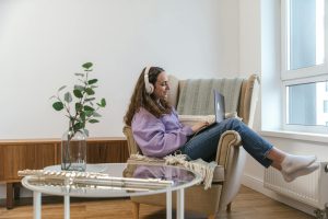 Young woman wearing headphones, smiling while working on a laptop at home, creating a cozy and relaxed atmosphere.